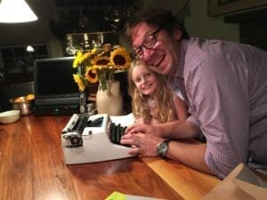 A man and young girl joyfully typing on a typewriter at a wooden table.